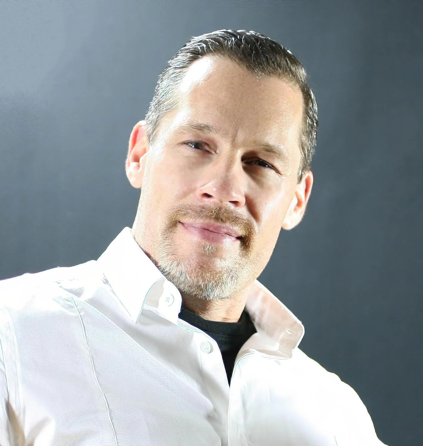 Close-up of a middle-aged man with short, dark, slicked-back hair, light skin, and a trimmed beard, wearing a white button-down shirt and a black undershirt, posing against a dark gray background.