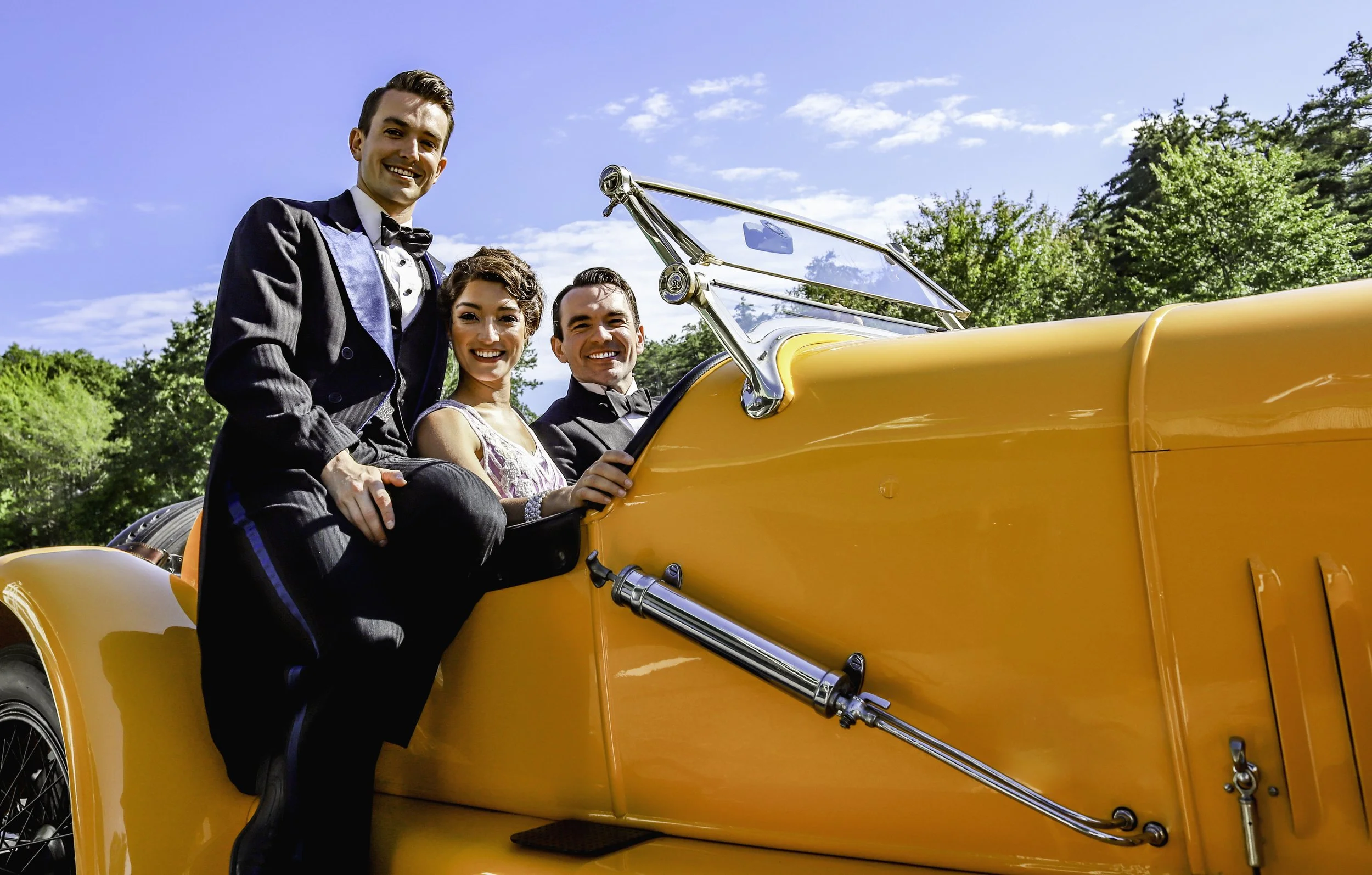 Three smiling people in formal attire sitting in a vintage yellow car outdoors on a sunny day with green trees and blue sky.