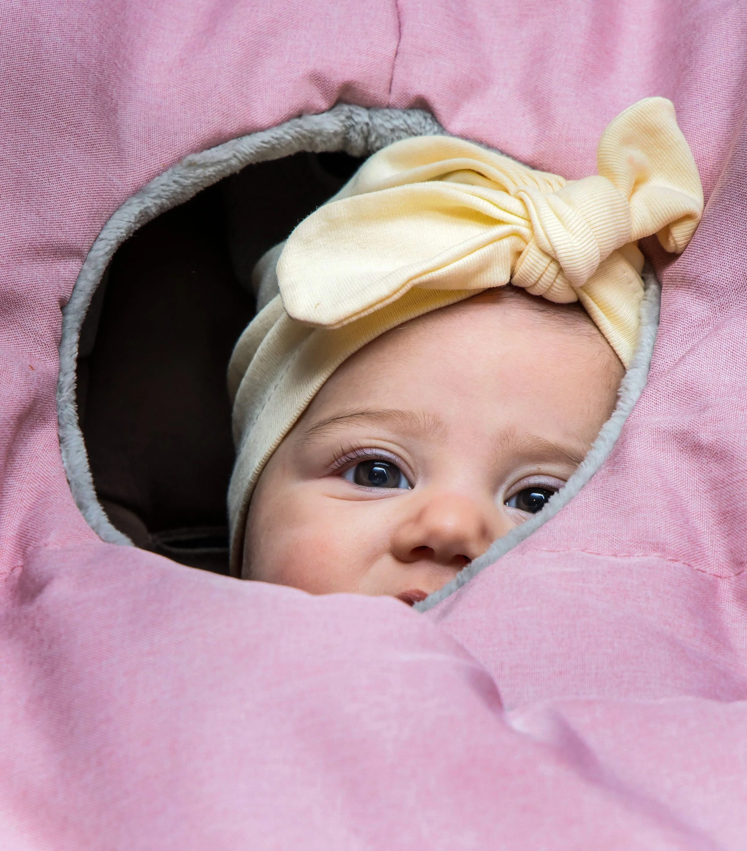 Close-up of a baby with brown eyes wearing a yellow headband with a bow, peeking out from a pink blanket.