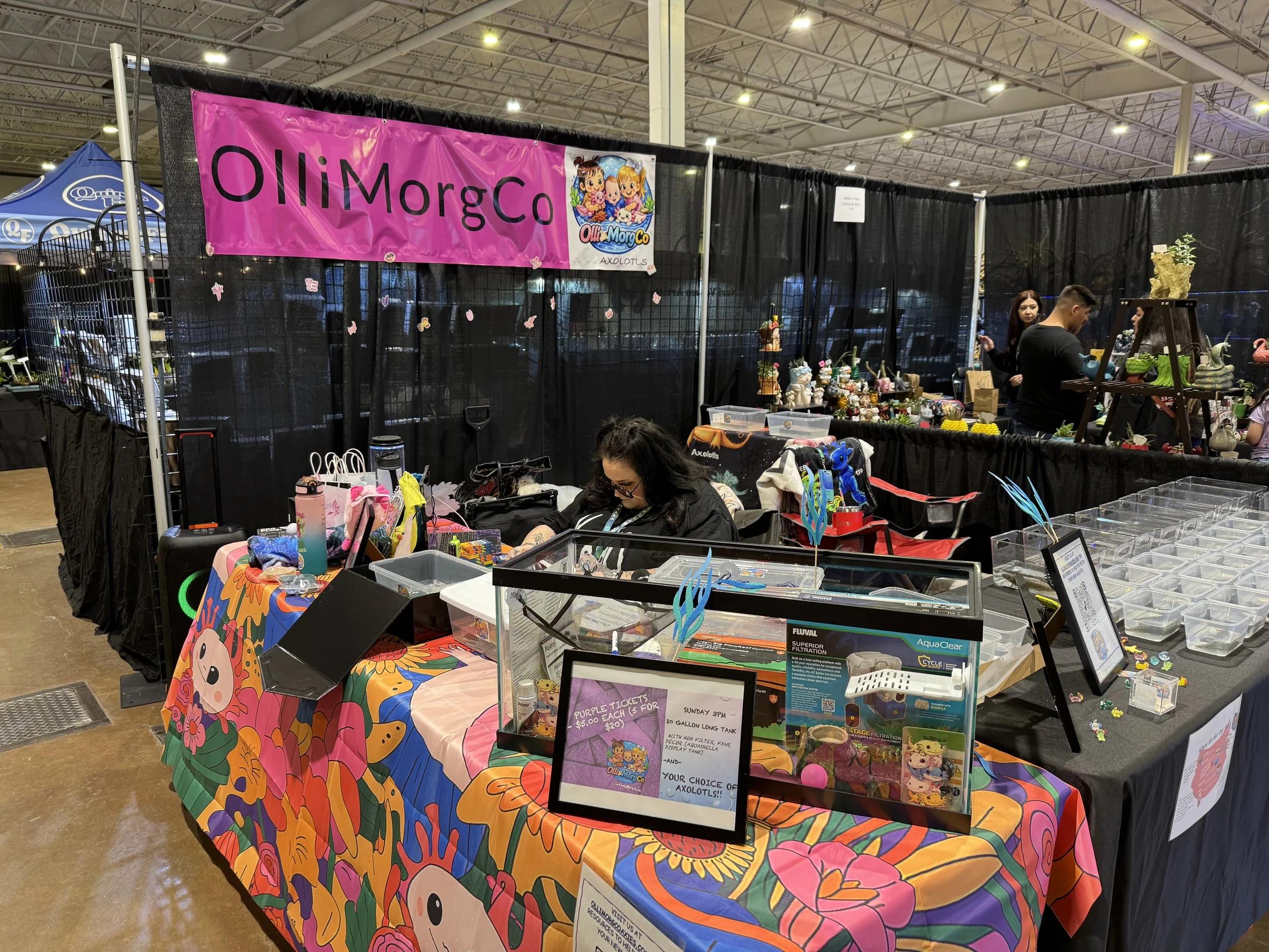 A booth at an indoor event displays colorful handmade crafts and figurines, with a pink banner reading 'OllieMorgCo,' featuring a cartoon logo of two kids with unicorns. Several people are browsing and working at the vendor table, surrounded by black curtains and various display items.