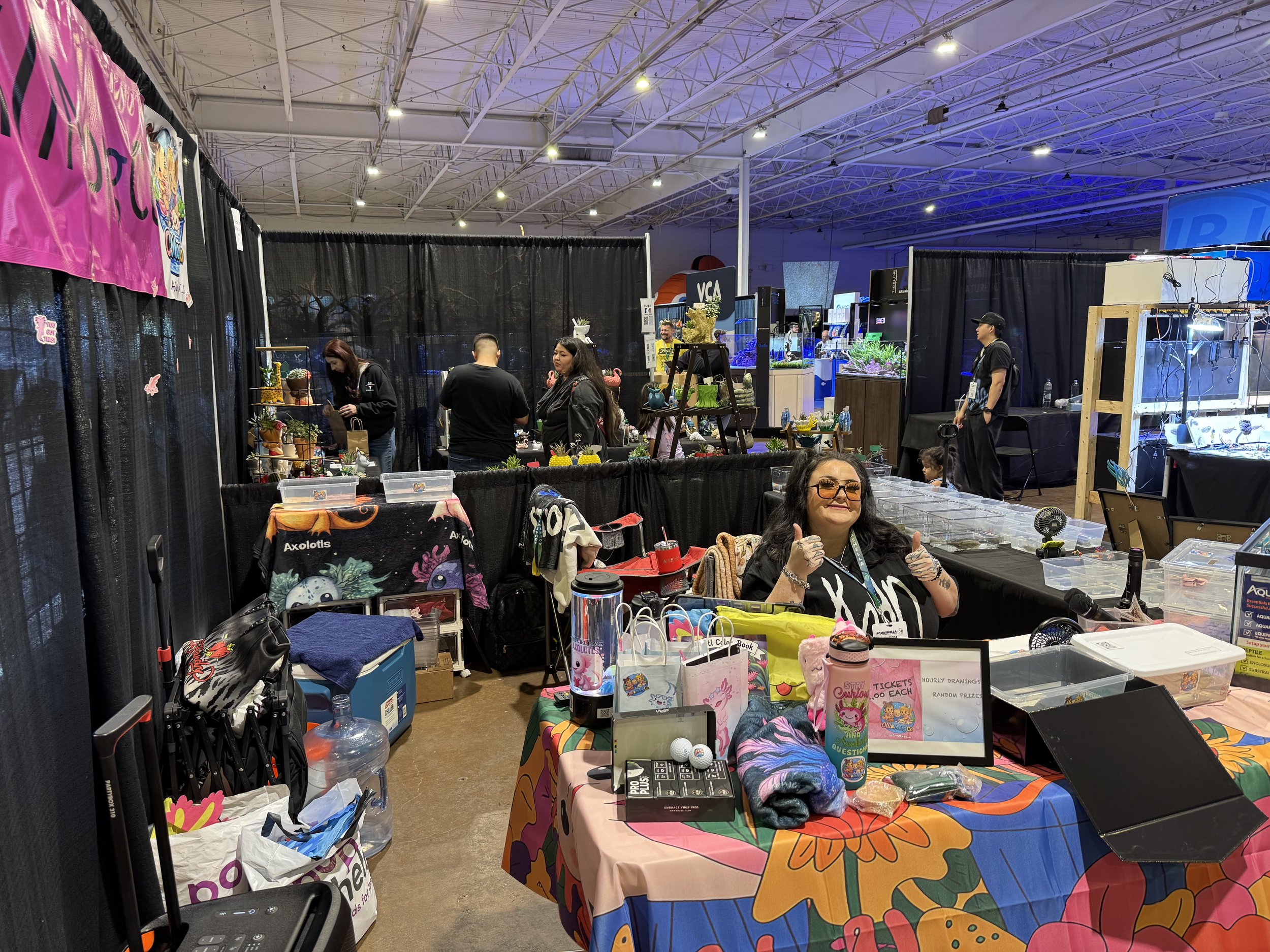 A scene inside an indoor event or convention with multiple vendor tables, black curtains, and people browsing and selling items such as plants, ceramics, and artwork.