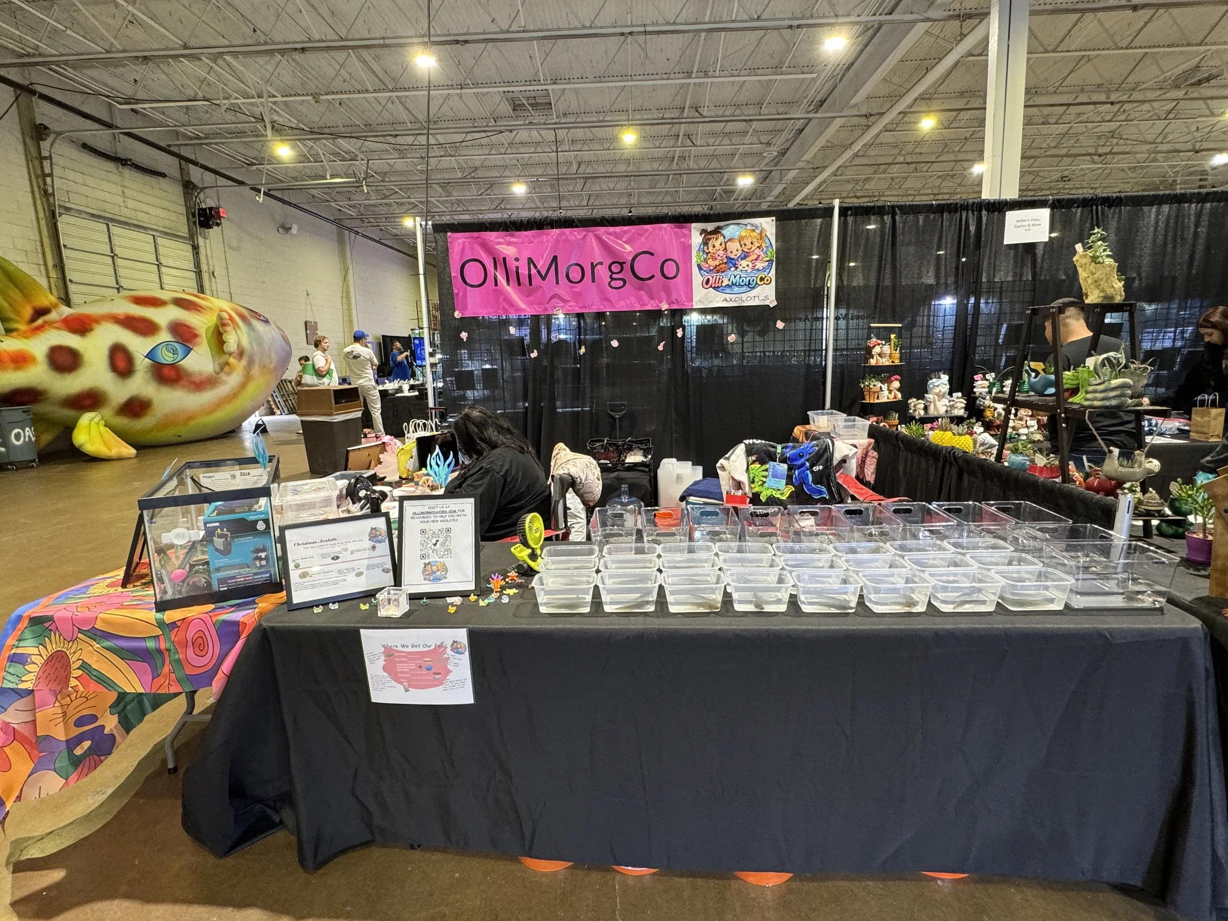 A convention booth for OlliMorgCo featuring various figurines, crafts, and a large fish sculpture in the background at an indoor event.