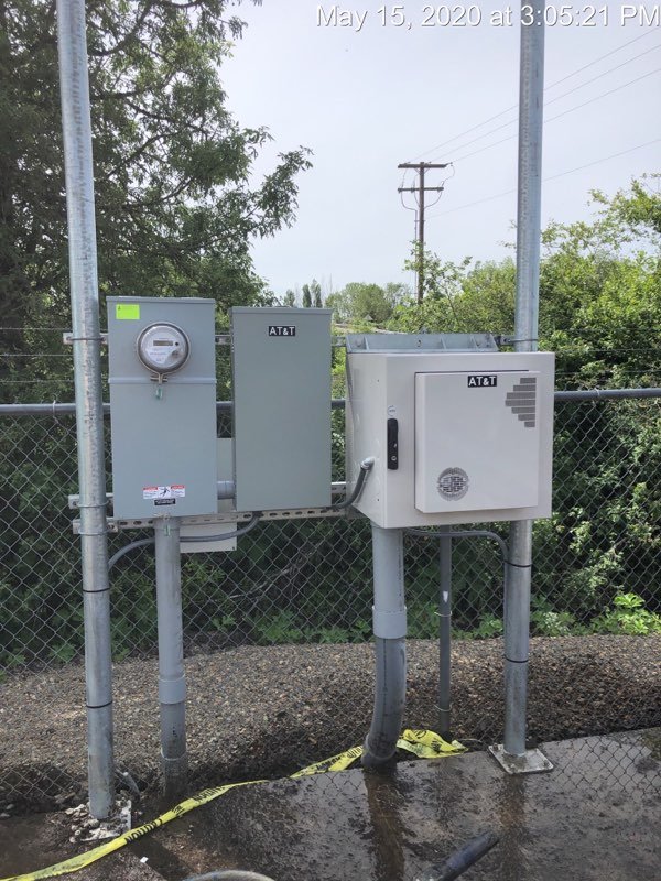 Three utility boxes mounted on a metal fence, with trees and power poles in the background, on a wet ground surface.