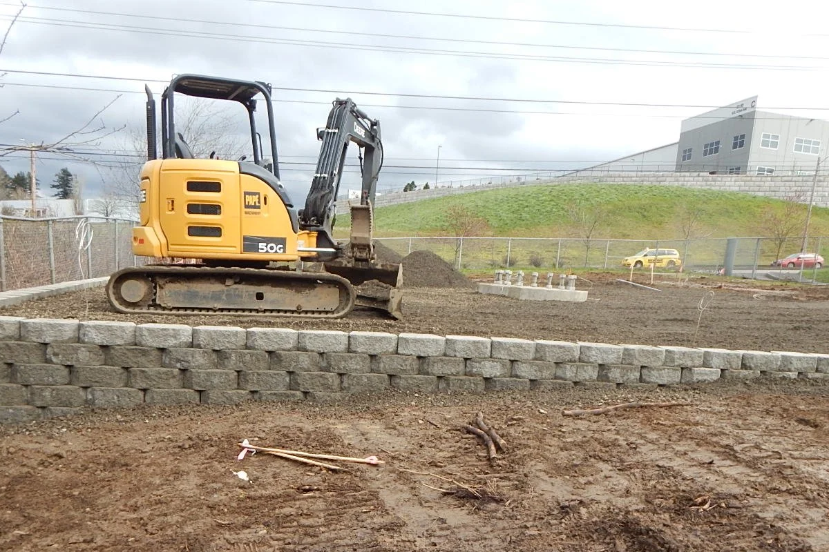 A small yellow excavator on a construction site, surrounded by dirt and a stone retaining wall, with a grassy hill, buildings, and parked cars in the background.