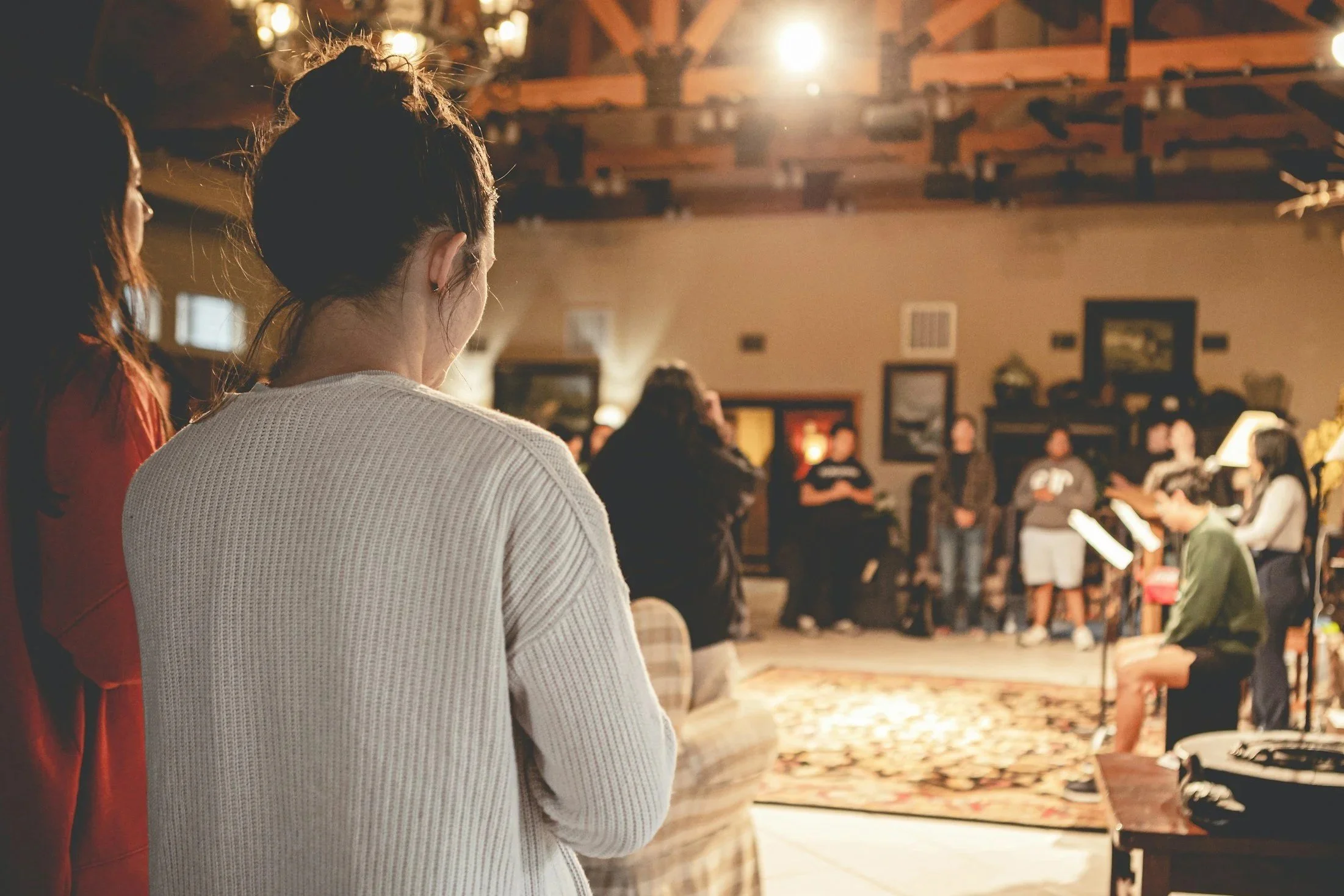 People gather in a warmly lit indoor space, possibly a community center or church, with some individuals singing or reading music and praying.