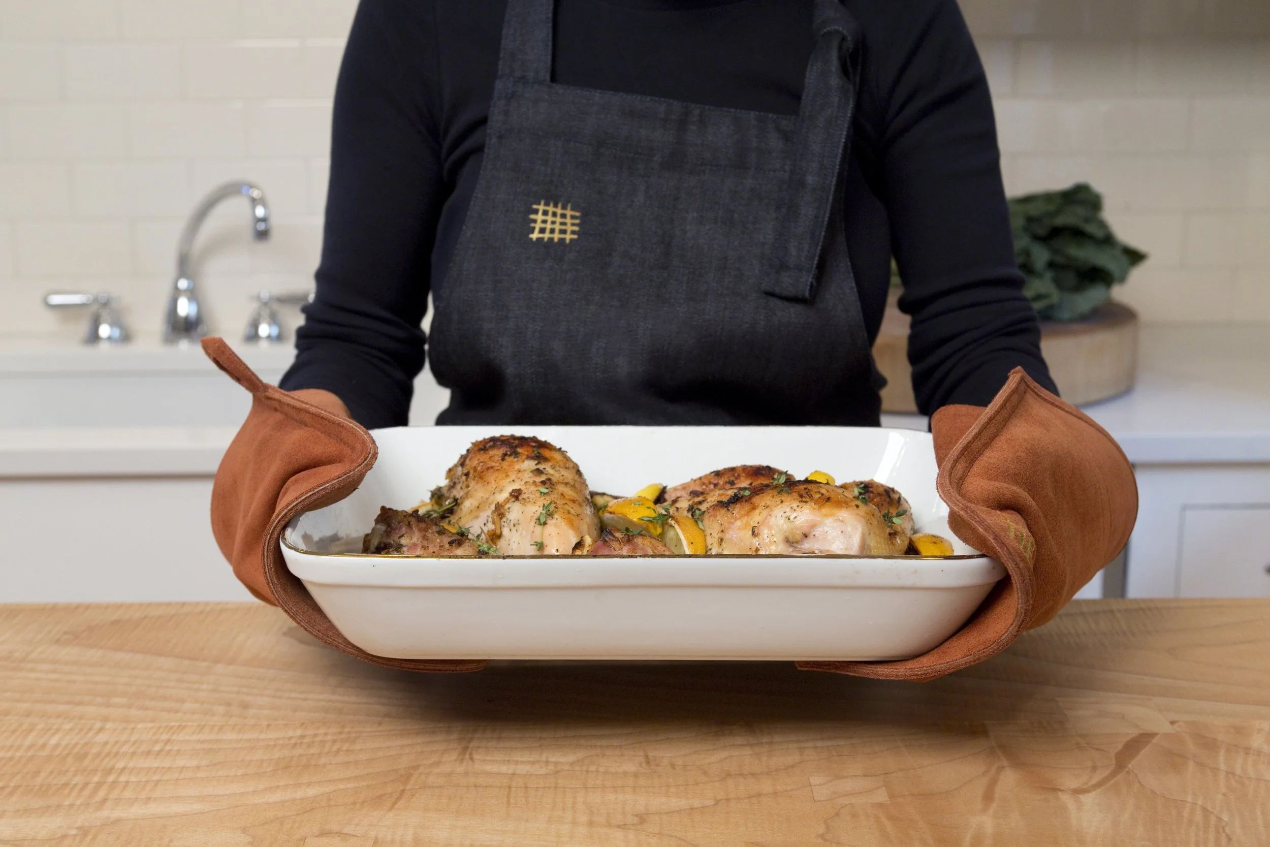 Person holding a white serving dish with cooked chicken and vegetables, wearing a dark apron and oven mitts, in a kitchen with a sink and leafy greens in the background.