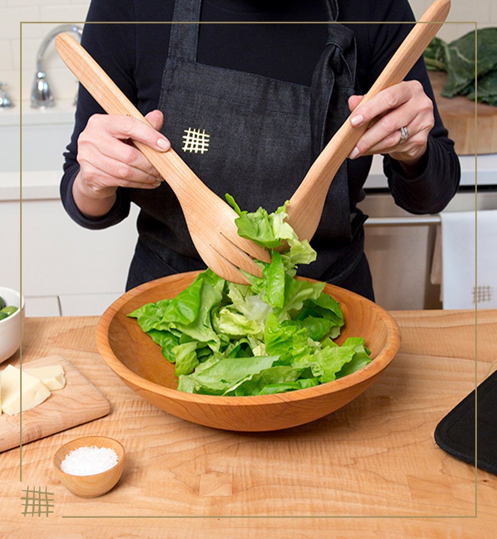 Person wearing a black apron mixing fresh green lettuce in a wooden bowl using two wooden salad servers in a kitchen.