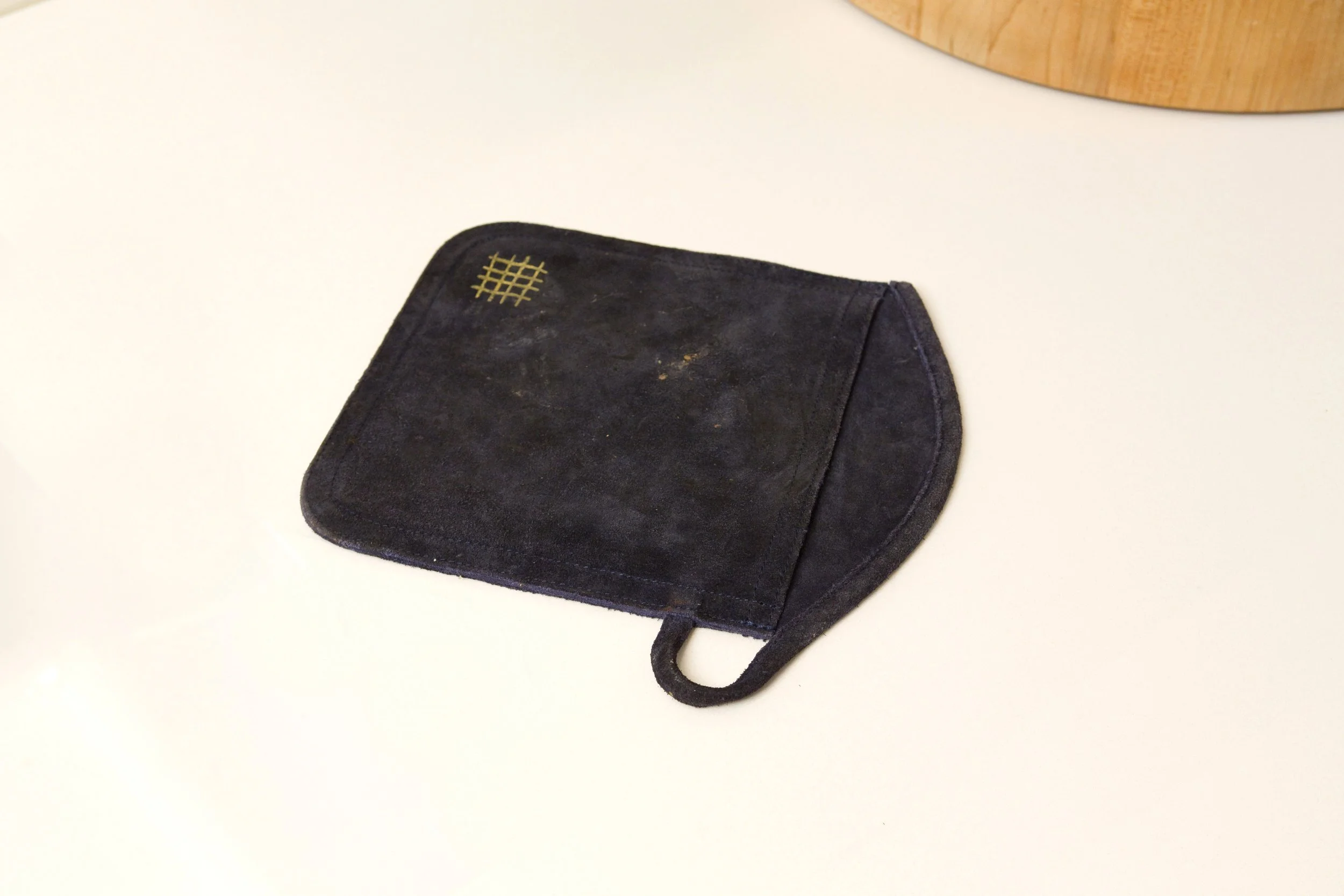A worn suede oven mitt with gold detailing on a white surface.