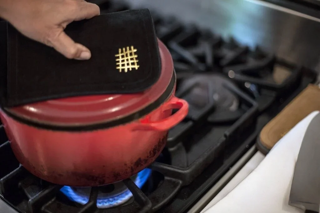 A red pot on a stovetop with a blue flame, and a person's hand holding a black oven mitt above the pot.