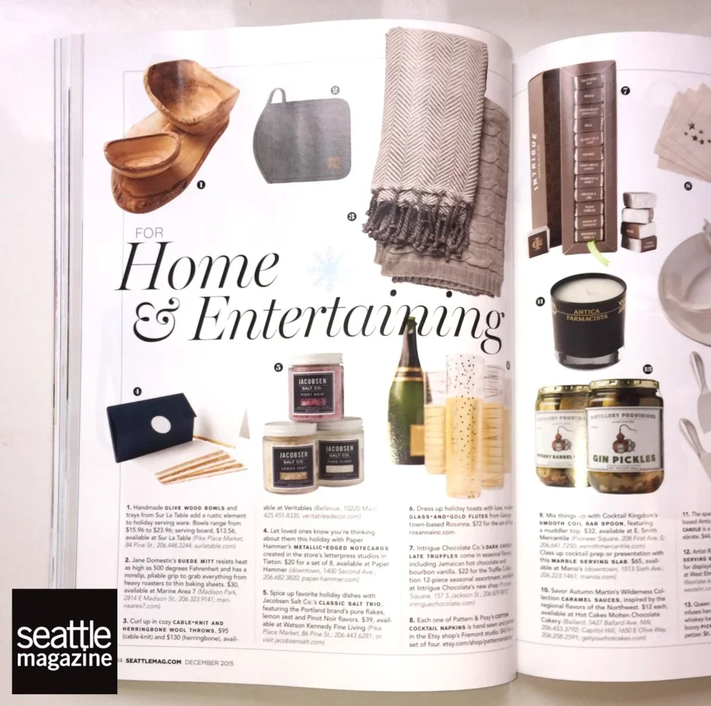 Magazine page showcasing home and entertaining items such as wooden bowls, a small gray bag, a blanket, a wooden tray, candles, jars of gherkins, chocolates, a candle, and decorative objects.