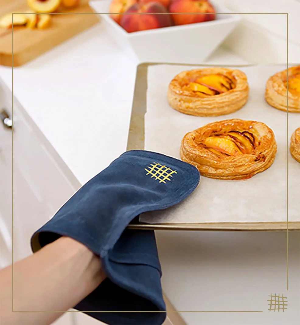 Person using an oven mitt to remove a tray of peach Danish pastries from an oven or baking sheet, with a bowl of peaches in the background.