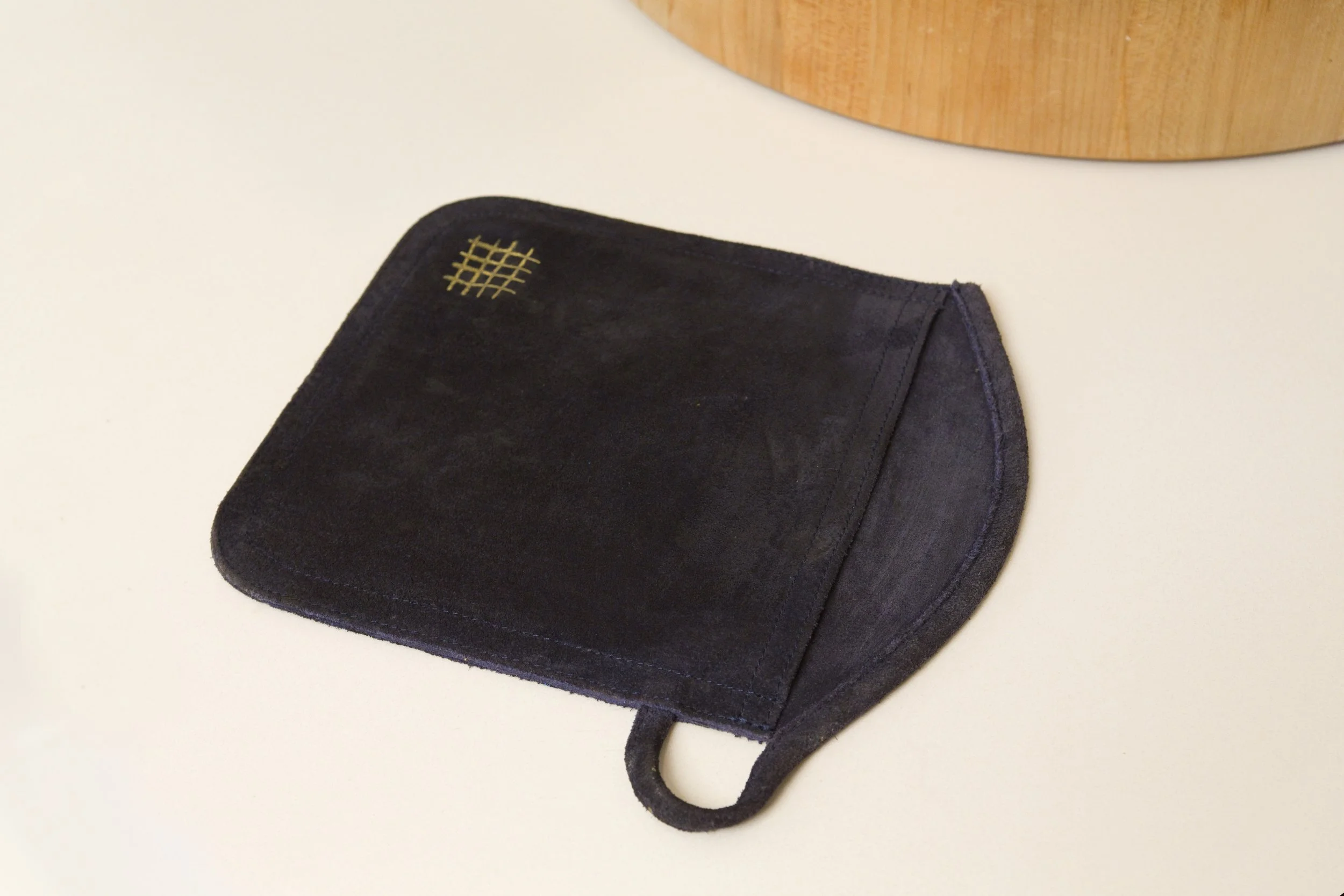 A suede oven mitt with a gold checkered design in the upper left corner, resting on a white surface near a wooden object.