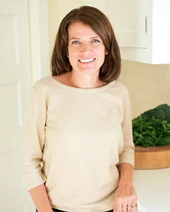 Woman with brown hair smiling in a light-colored sweater, standing in a kitchen with a white cabinet and a green plant in the background.