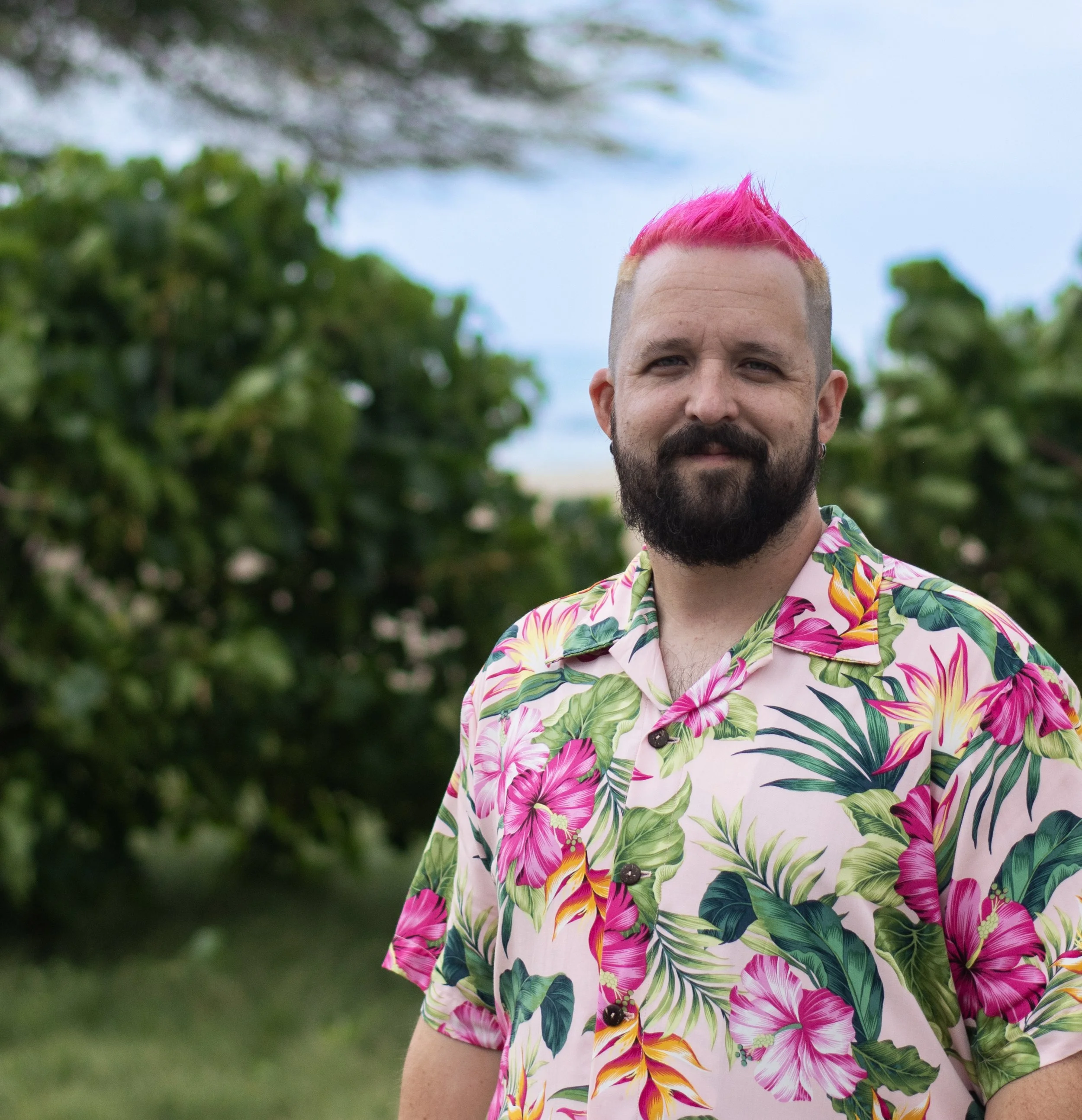 A man with a pink Mohawk, a beard, and earrings, wearing a colorful tropical shirt, standing outdoors with greenery and trees in the background.