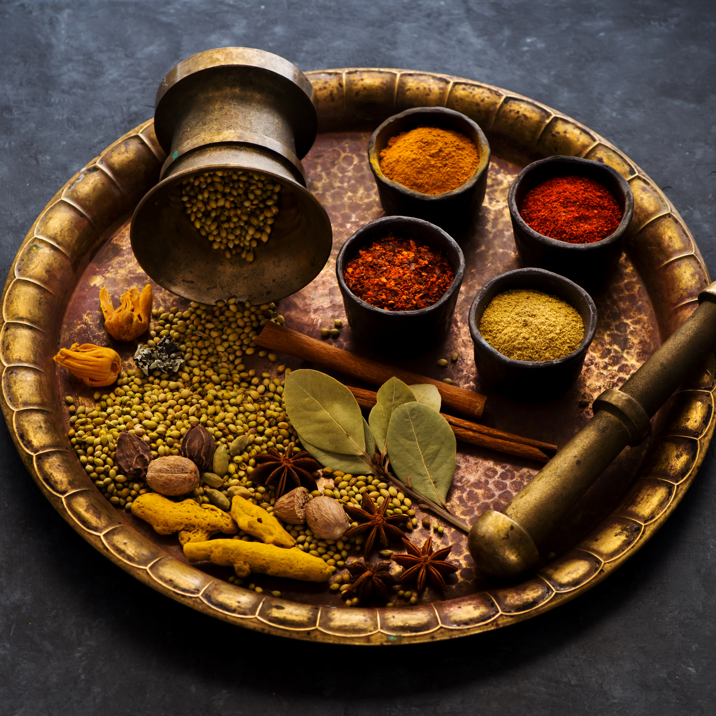 A brass tray with various Indian spices, dried herbs, and a mortar and pestle on a dark surface.
