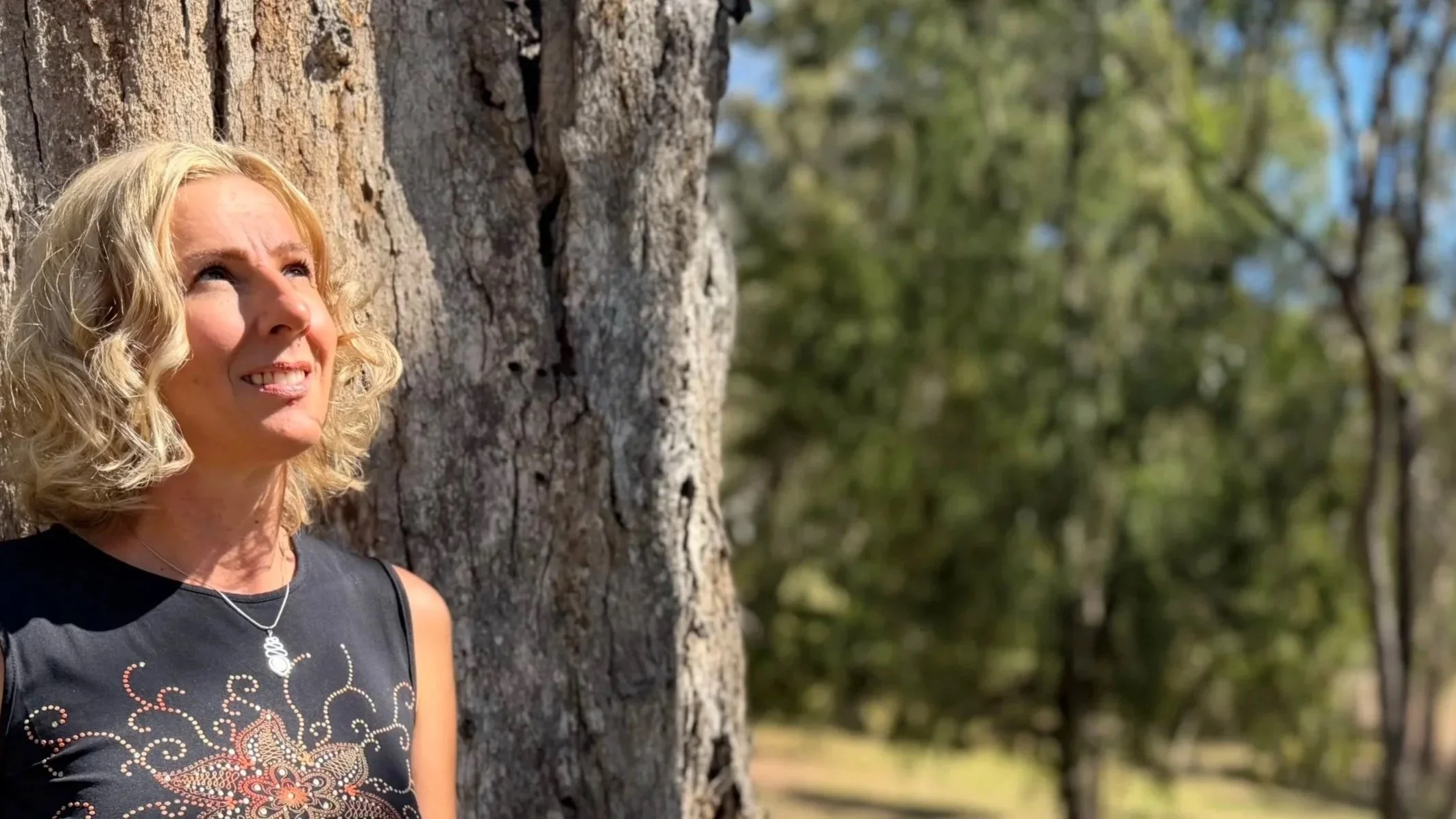 A woman with curly blonde hair smiling while leaning against a tree outdoors with green foliage in the background.