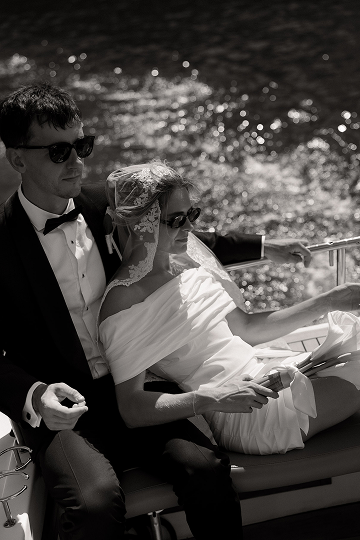 A couple in wedding attire riding on a boat, with water in the background.