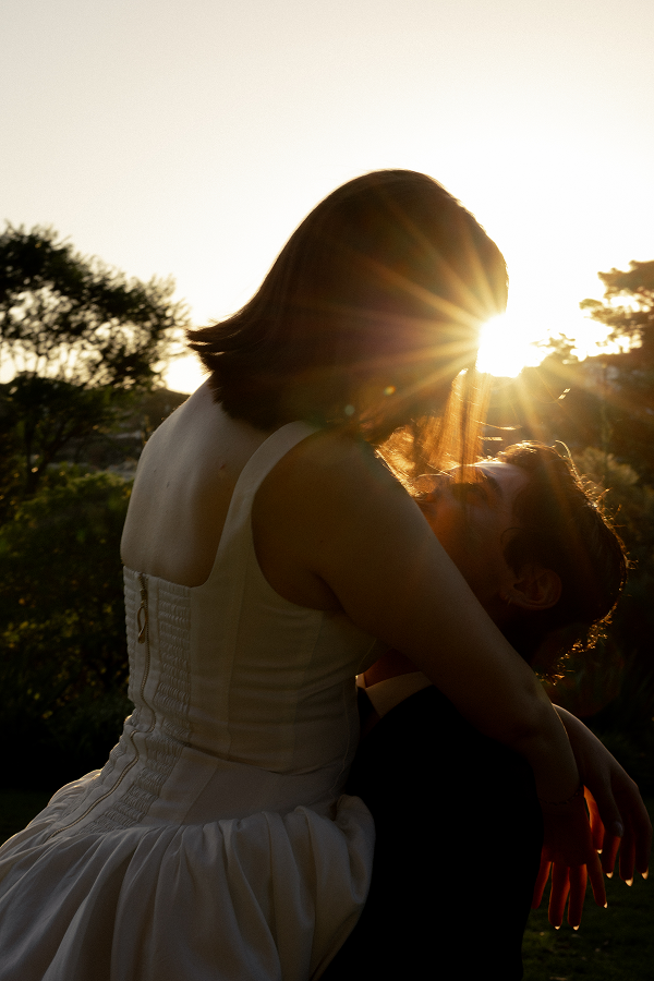 A couple outdoors during sunset, with the woman wearing a white dress and the man in dark clothing, embracing affectionately.