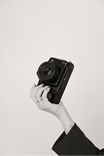 Person holding a camera with a strap, photographed against a plain background.