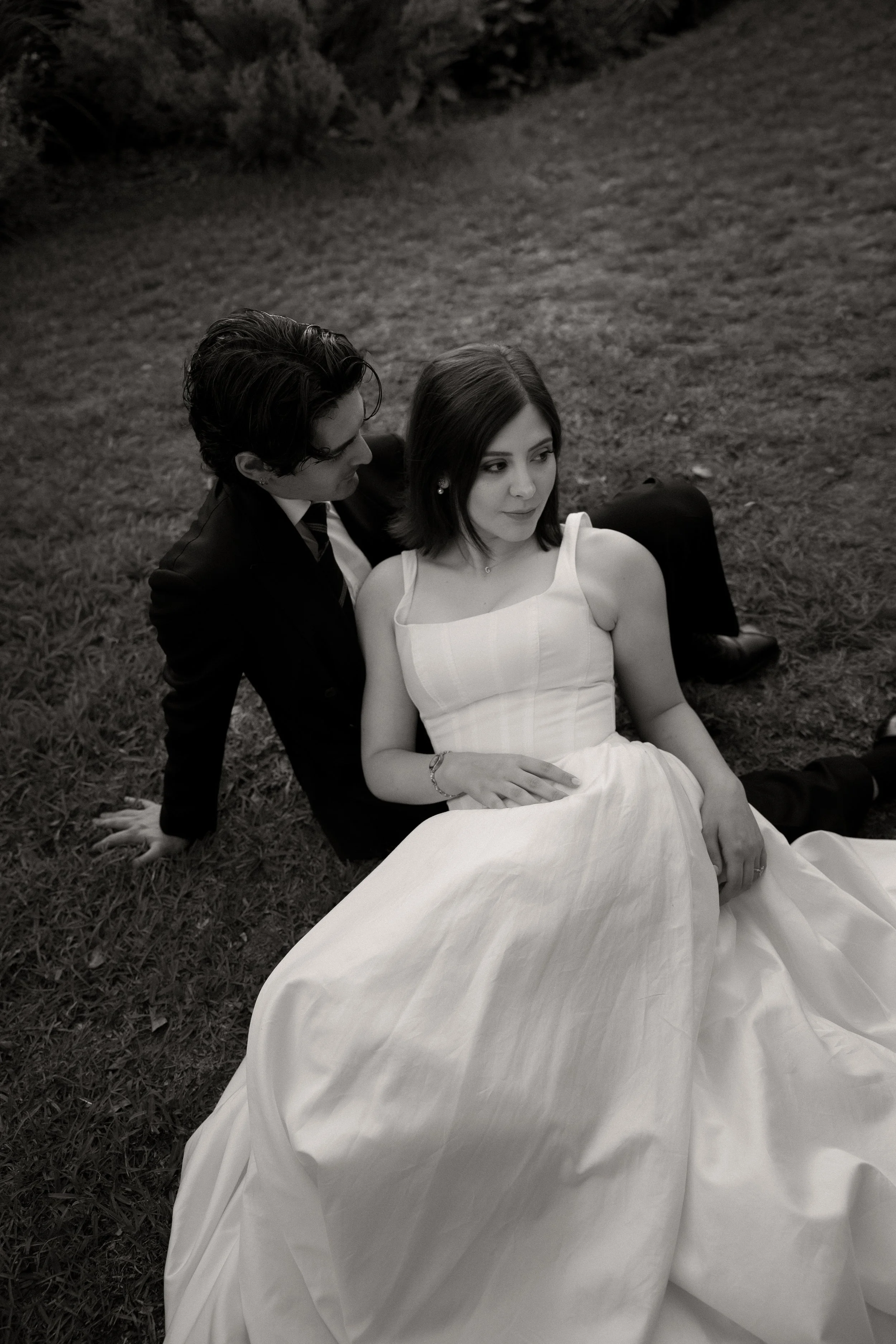 A black and white photo of a young couple sitting on the grass outdoors. The man, dressed in a suit, is sitting behind the woman, who is wearing a white dress, and they appear to be in a romantic moment.