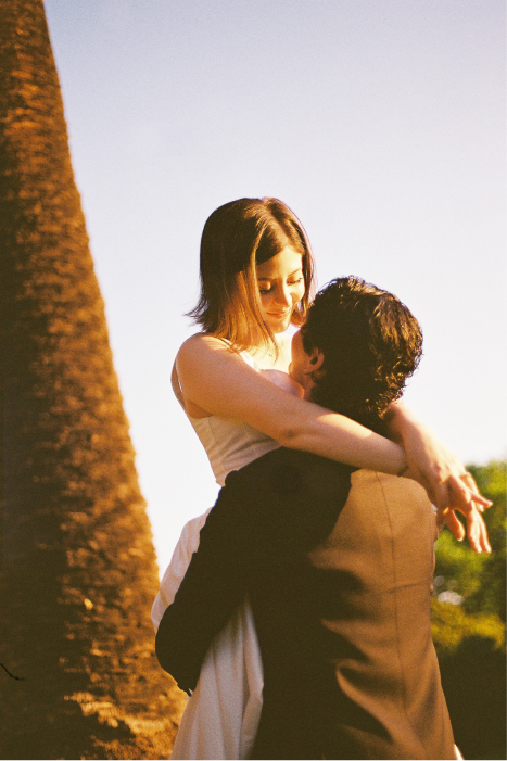 A couple embracing outdoors during sunset, with the woman in a white dress and the man in a black suit, beside a large tree and clear sky.