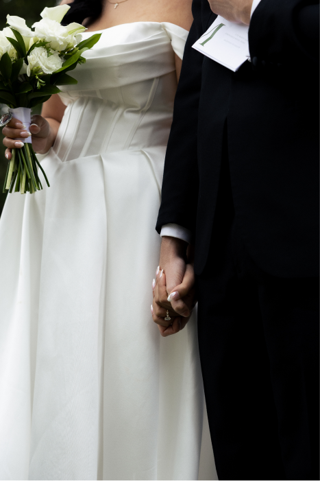 Close-up of a bride and groom holding hands during a wedding ceremony, the bride in a white dress holding a bouquet of white flowers, the groom in a black suit.