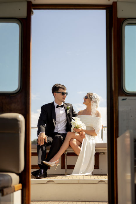 A newlywed couple sitting on a boat deck, dressed in wedding attire, with a bright blue sky in the background.