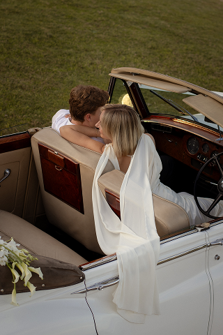 A couple sharing a kiss in a vintage white convertible car on grass.