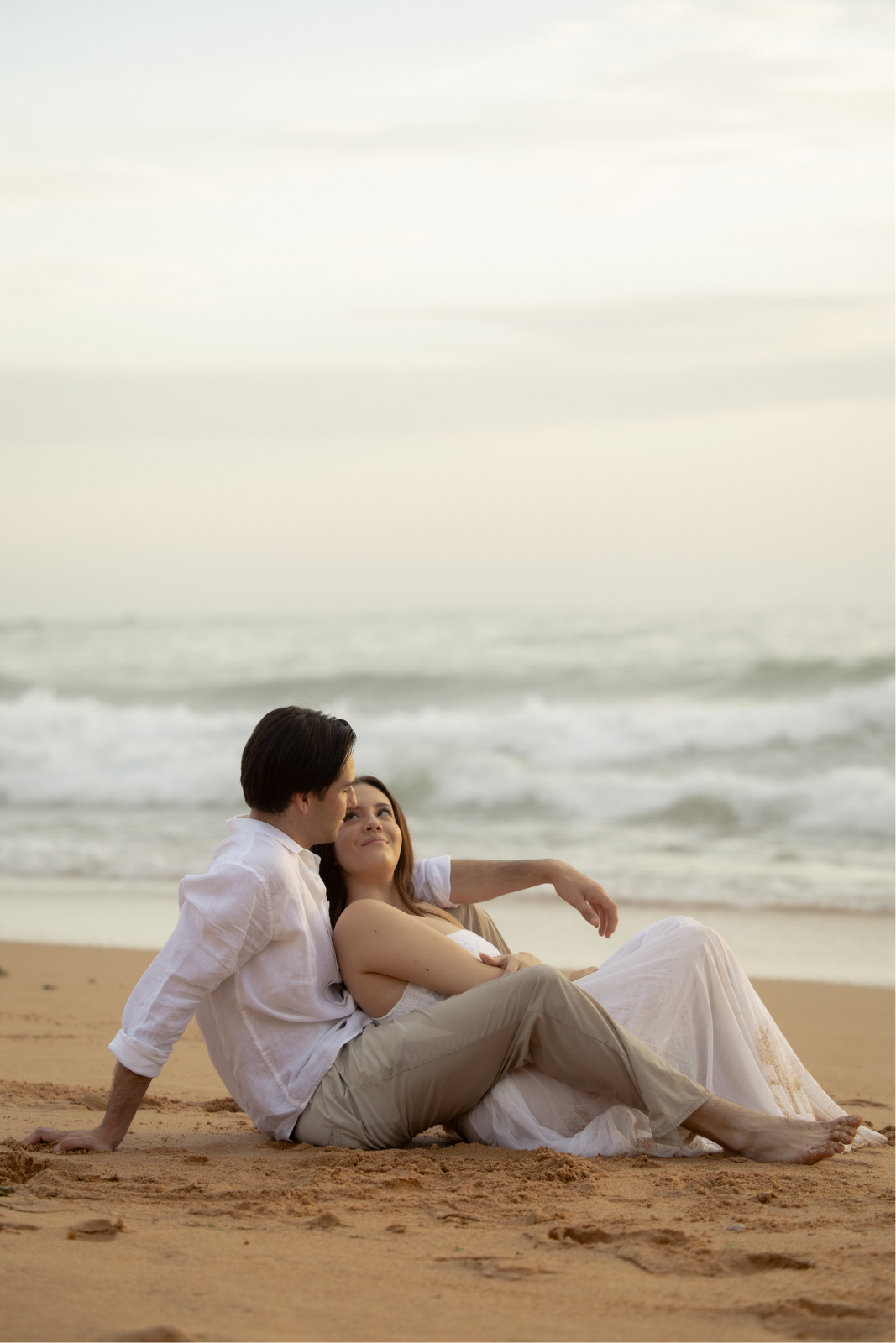 A couple sitting on the beach, facing each other, with ocean waves in the background.