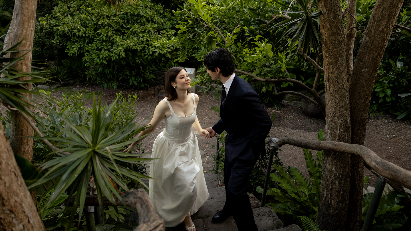 A woman in a white dress and a man in a black suit holding hands and looking at each other on a wooded pathway surrounded by lush green plants and trees.