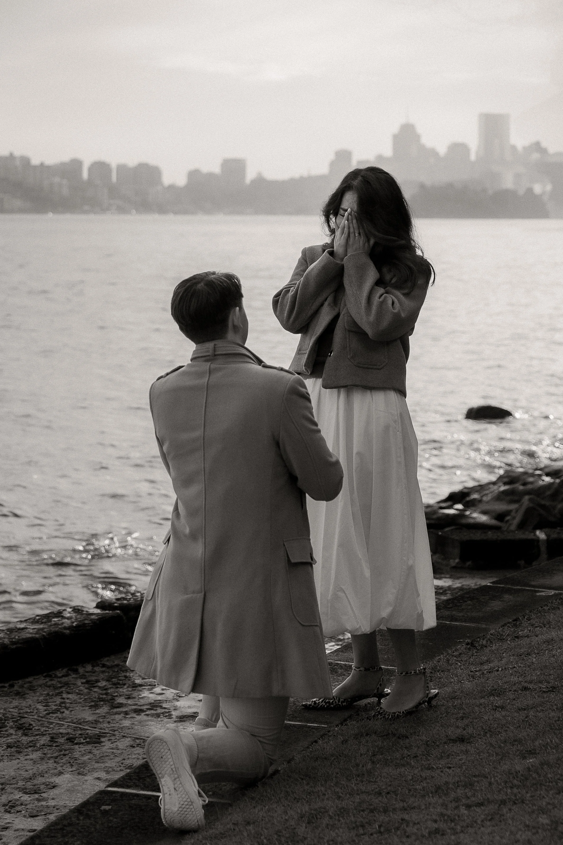A woman is standing by a body of water with a city skyline in the background. She is covering her face with her hands. A man is kneeling on a skateboard, facing her. The scene is in black and white.