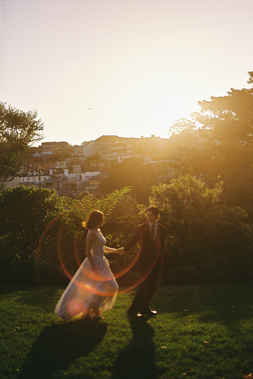 Couple dancing outdoors on grass with sunset in the background, trees, and houses on a hillside.