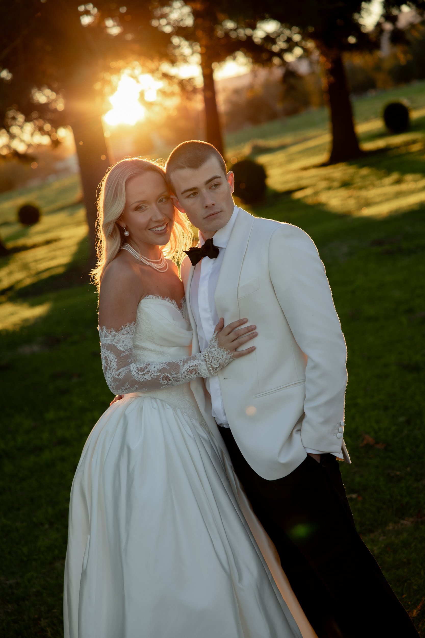 A bride and groom standing close together outdoors at sunset, with trees and grass in the background. The bride is smiling, wearing a white wedding dress with lace details, and pearls. The groom, wearing a white tuxedo jacket and black bow tie, has his arm around her. The sunlight creates a warm glow behind them.