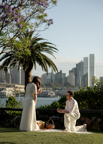 A man proposing marriage to a woman in a park near a city skyline, with trees and flowers in the background.