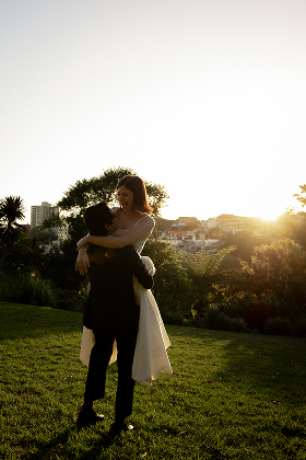 A couple dancing outdoors at sunset in a park with city buildings in the background.