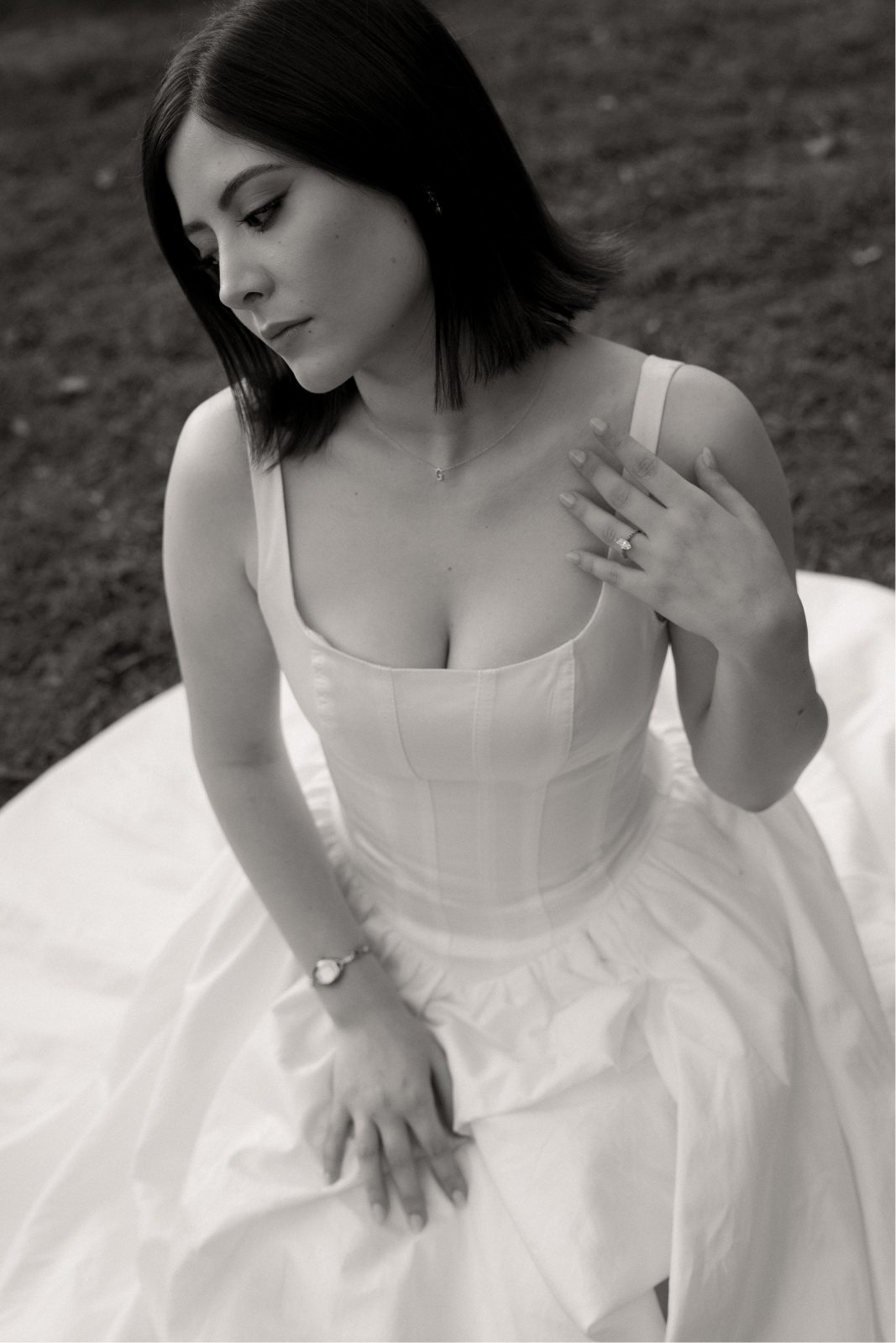 Black-and-white photo of a young woman in a sleeveless dress, sitting outdoors on the ground with a thoughtful expression, her left hand resting on her chest and wearing a ring and watch