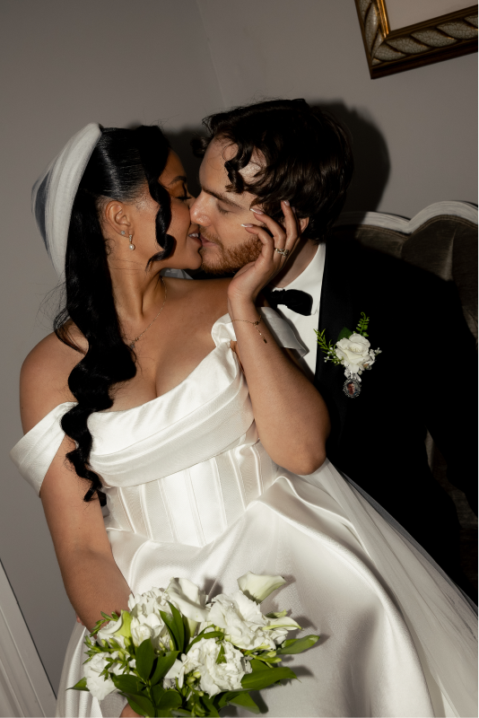 Bride and groom sharing a kiss, bride holding a bouquet of white flowers, bride wearing a white wedding dress and veil, groom in a black tuxedo with bow tie, indoor setting with a framed picture on the wall.