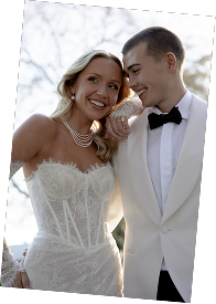 A bride and groom smiling and looking at each other, outside during daytime, bride wearing a white wedding dress and pearl necklace, groom in a white tuxedo with black bow tie.
