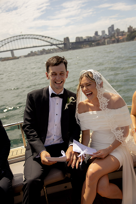 A newlywed couple on a boat, smiling and holding a paper, with the Sydney Harbour Bridge in the background.
