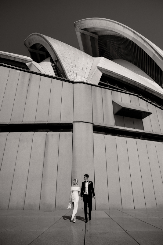 A couple walking in front of a modern, architecturally distinctive building with large, curved structures, captured in black and white.