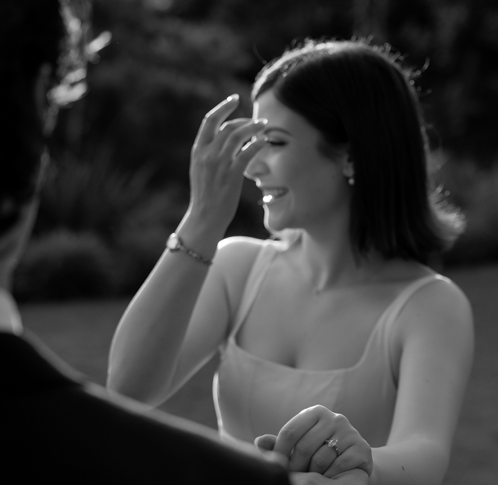A woman smiling and laughing outdoors, wearing a sleeveless top, with a man whose face is not visible.