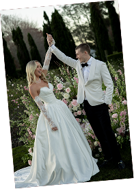 A bride and groom dancing outdoors at their wedding, holding hands up in the air with garden and flowers in the background.