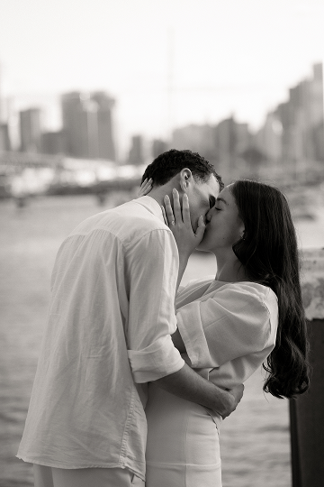 A couple sharing a kiss outdoors near a city skyline in black and white.