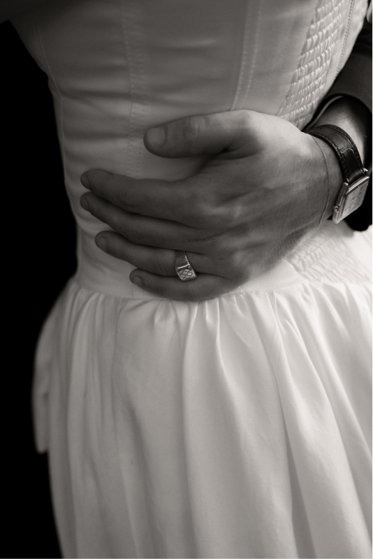 Close-up of a person's hand resting on their midsection, wearing a wedding dress, a ring, and a wristwatch.