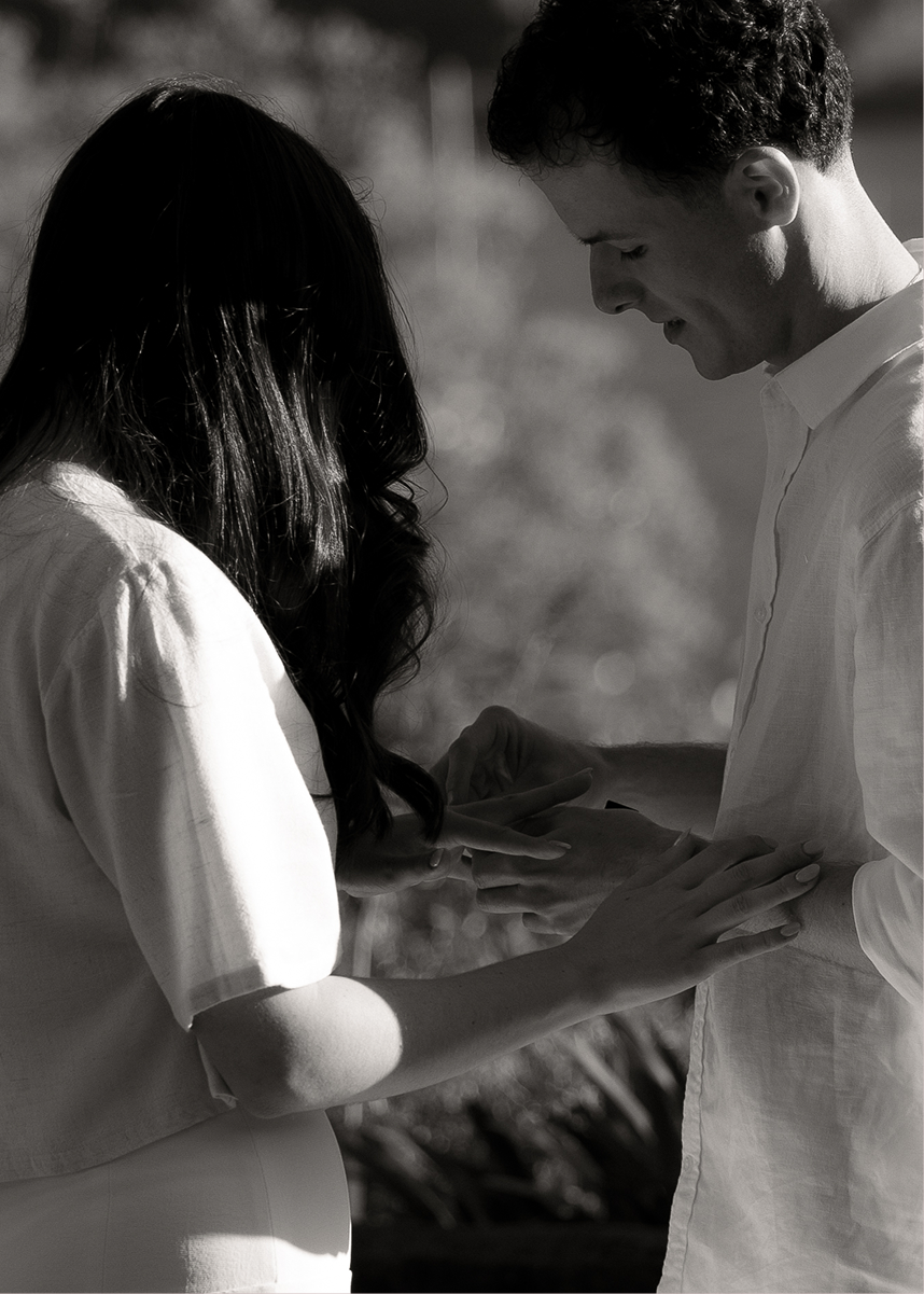 A man and woman exchanging rings outdoors, with the woman placing a ring on the man's finger, in black and white.