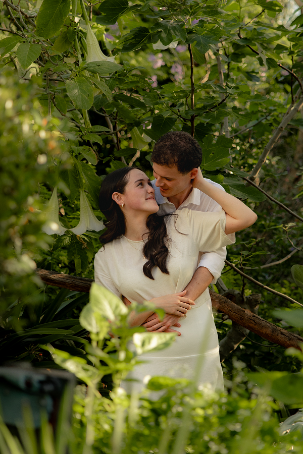 A young couple embracing in a lush garden filled with green leaves and large, white trumpet-shaped flowers.
