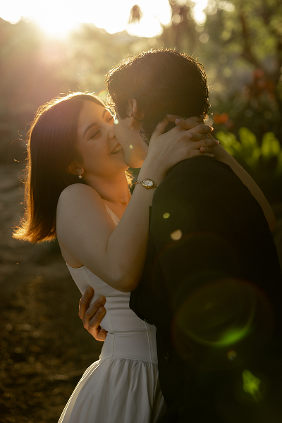 A couple embraces and kisses outdoors during sunset, with sunlight creating a warm glow around them.