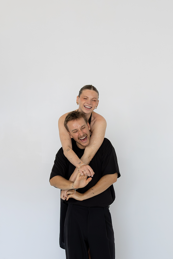 A woman giving a piggyback ride to a man, both smiling and laughing against a plain white background.