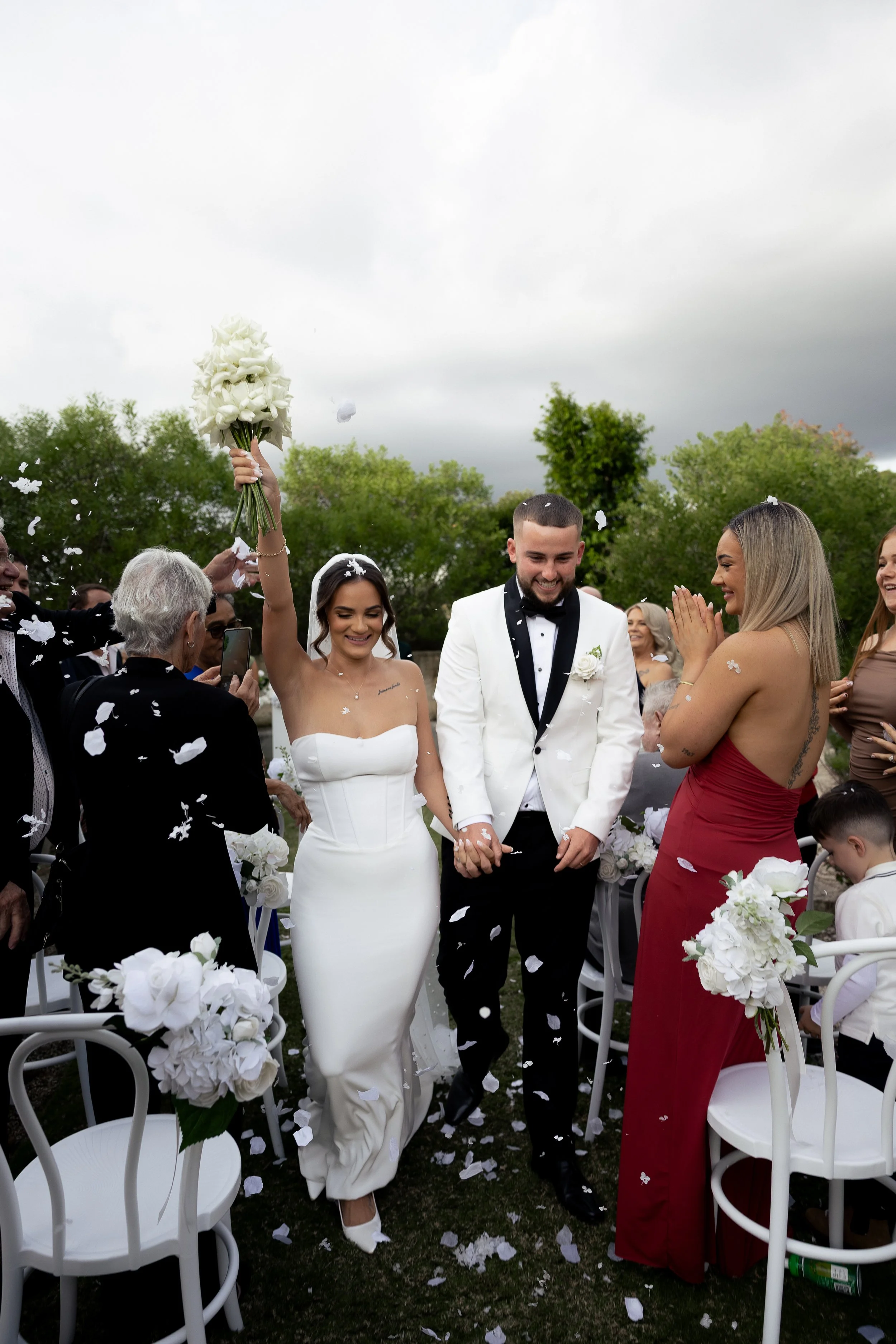 A bride and groom walk back down the aisle immediately after their ceremony. The bride is in a white dress and veil and has her bouquet of flowers lifted above her head in celebration. The groom wears a white tux and is smiling with joy. 