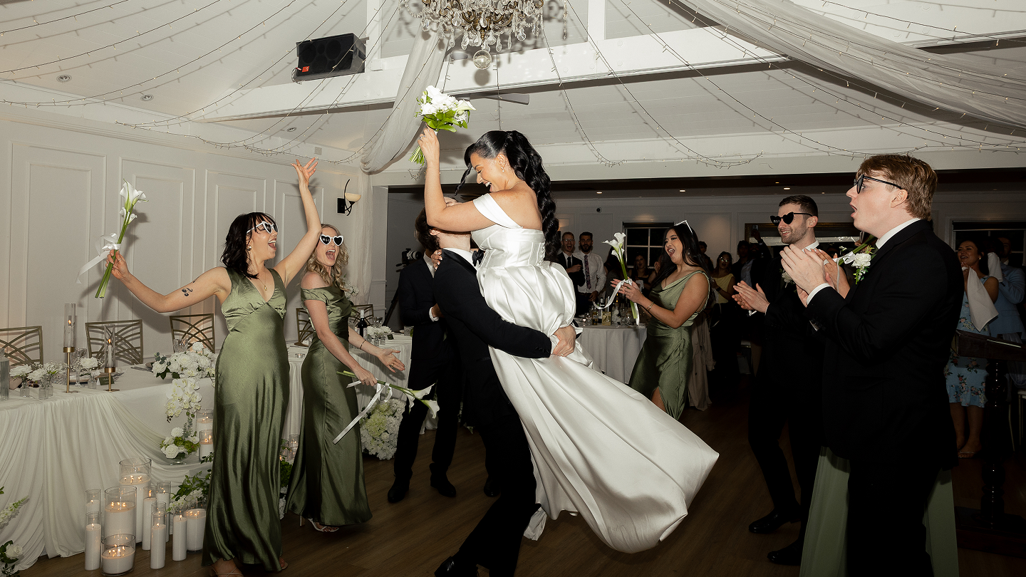 Bride in white dress being lifted by groom in tuxedo at wedding reception, surrounded by bridesmaids in green dresses and groomsmen, all celebrating indoors with decorations and candles.